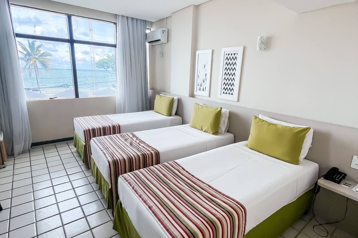 A hotel room with three beds, a window with a view of the sea, and a tiled floor.