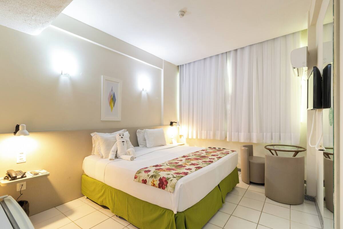 A hotel room with a bed, bedside lamps, a telephone, a mirror, a small table, and a window with curtains.