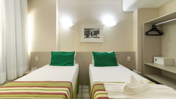 A hotel room with two single beds, a green pillow on each, a framed picture on the wall, a white towel on the bed, and a white hanging garment on the right. - Hotel Golden Park Recife By Nacional Inn (Recife)