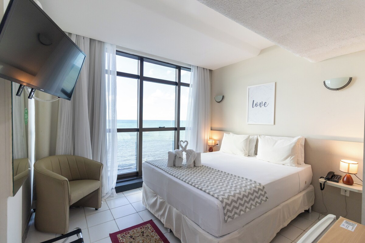 Habitación de hotel con cama, sillón, televisión, ventana con vistas, mesita de noche con lámpara y un cartel en la pared que dice 'love'.