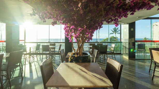 A restaurant with a view of the ocean, tables, chairs, and a large potted plant with pink flowers. - Hotel Golden Park Recife By Nacional Inn (Recife)