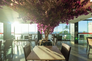 Un restaurante con vista al océano, mesas, sillas y una planta grande en maceta con flores rosadas.