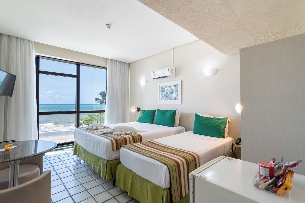 Habitación de hotel con dos camas, un escritorio, una silla, televisión y vista a la playa.