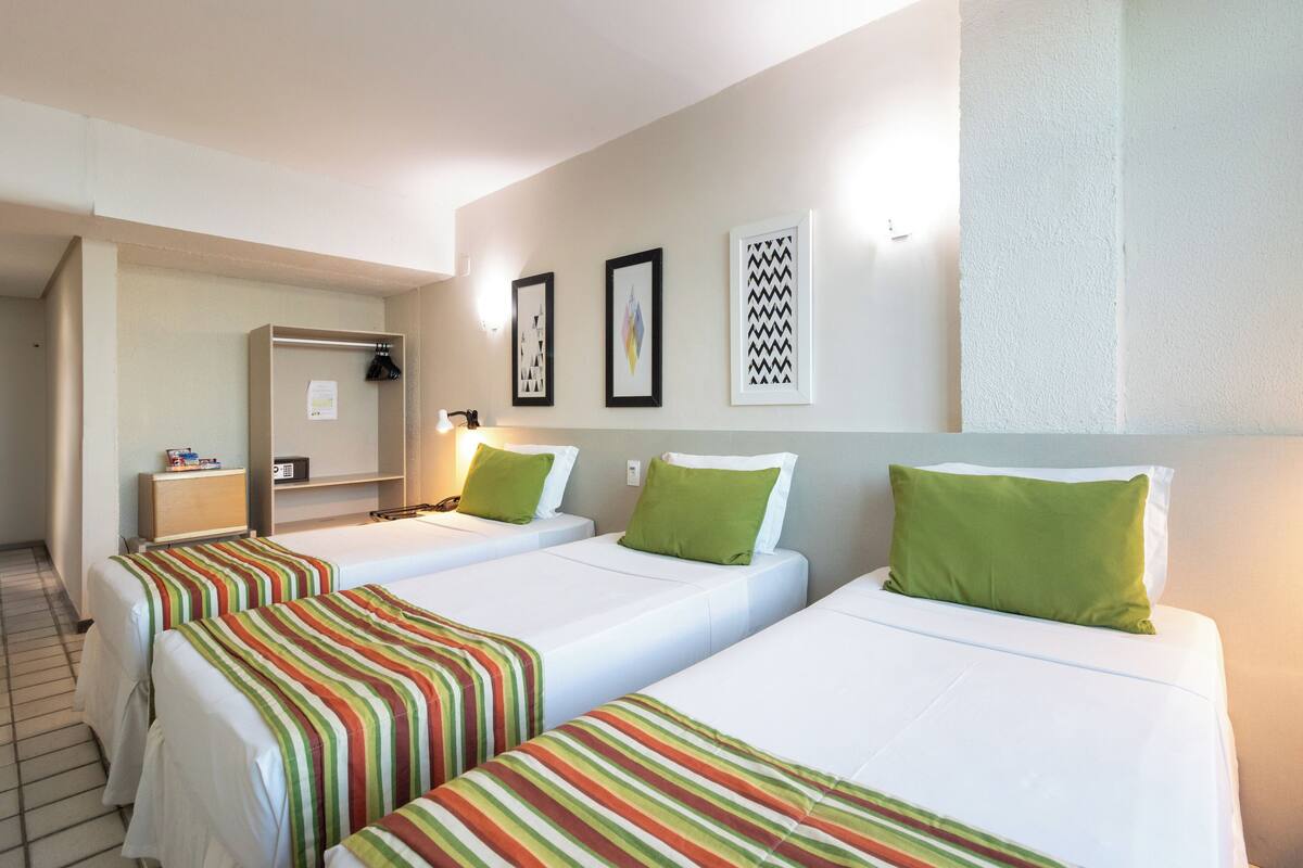 A hotel room with three single beds, each with a striped bedspread and a green pillow. There is a desk with a lamp and a telephone. The room has a white wall with framed artwork and a textured accent wall.
