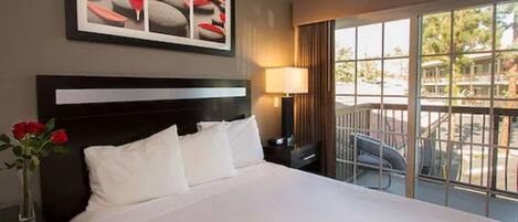 A hotel room with a large bed, a nightstand, a lamp, a vase with red roses, and a window with a view of the outdoors.