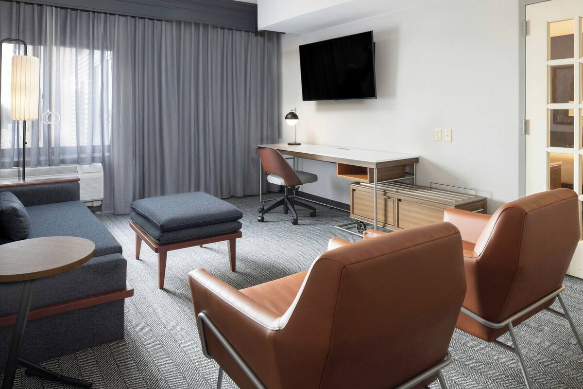 A hotel room with a grey sofa, two brown leather chairs, a small round table, a desk with a chair, a TV mounted on the wall, and a window with grey curtains.