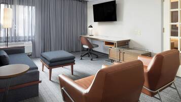 Ein Hotelzimmer mit grauem Sofa, zwei braunen Ledersesseln, einem kleinen runden Tisch, einem Schreibtisch mit Stuhl, einem an der Wand befestigten Fernseher und einem Fenster mit grauen Vorhängen.