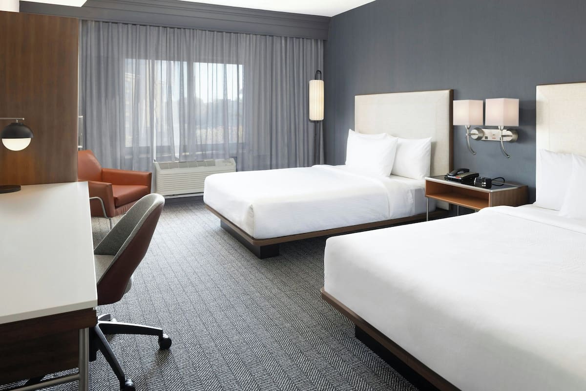 A hotel room with two beds, a desk, a chair, a lamp, and a nightstand with a phone and lamp.