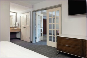 Een hotelkamer met een bed, een kaptafel, een spiegel, een wastafel en een deur naar een andere kamer.