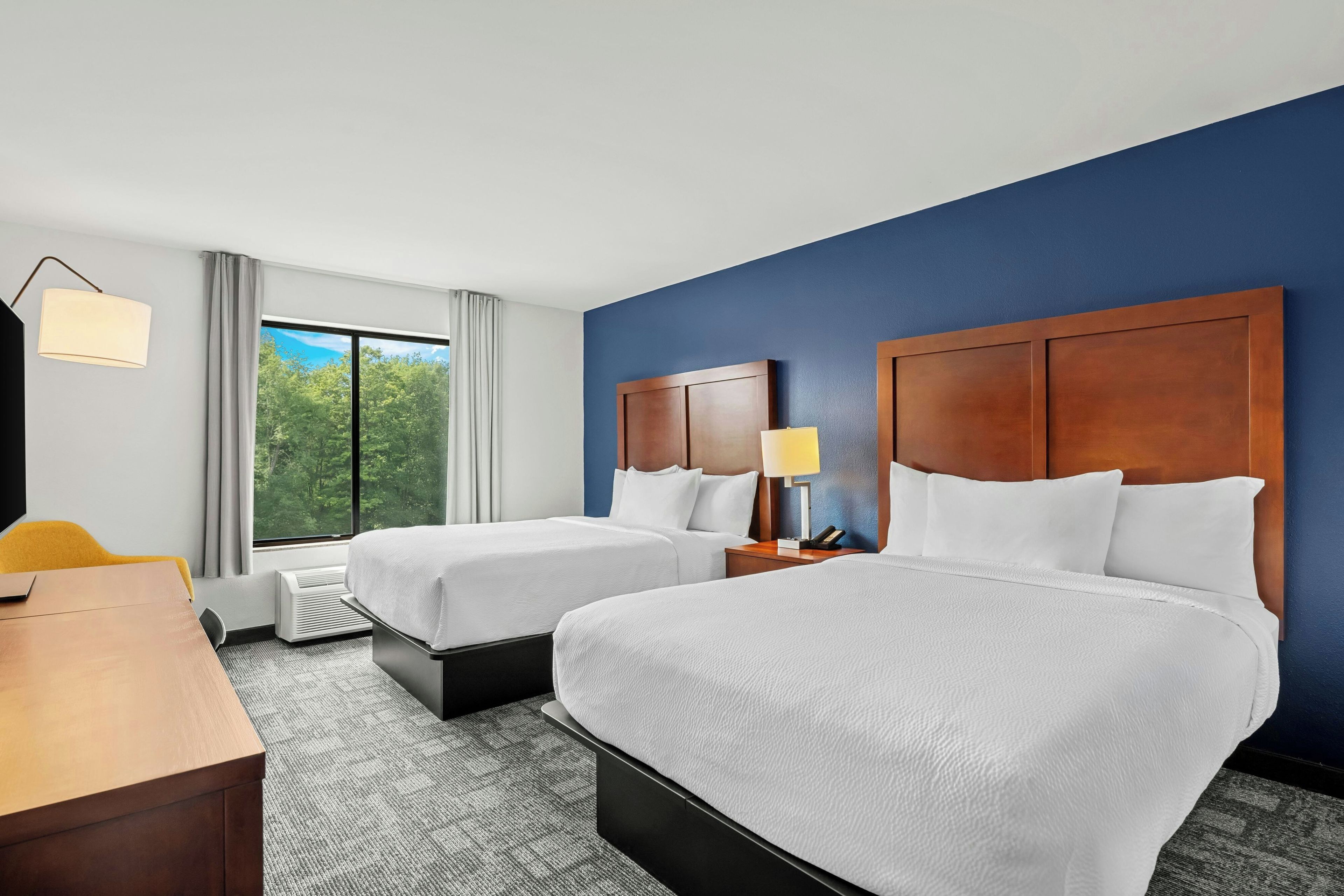 A hotel room with two beds, a desk, a chair, a lamp, and a view of trees through the window.