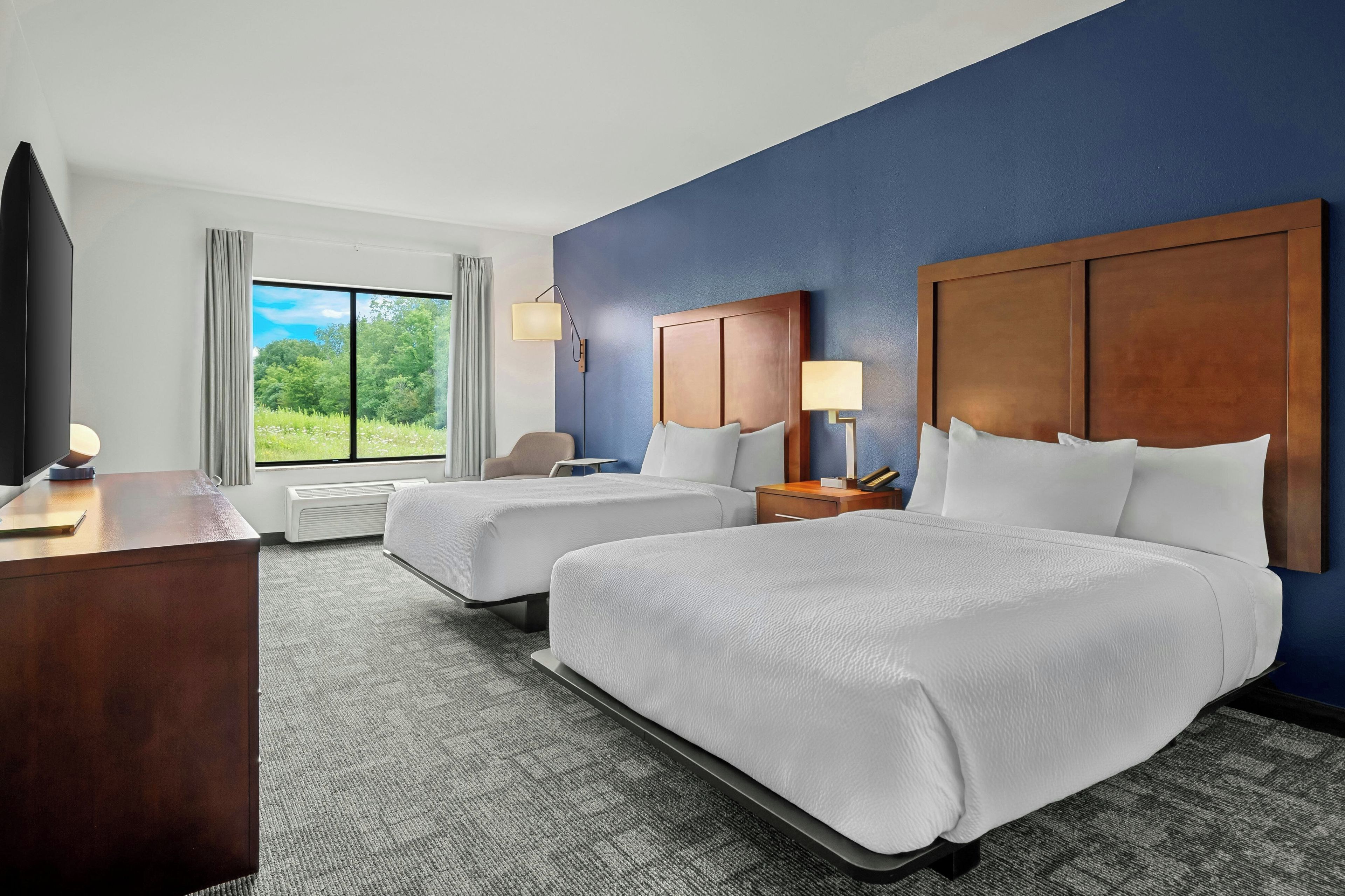 A hotel room with two beds, a desk, a chair, a window with a view of greenery, and a wall-mounted lamp.