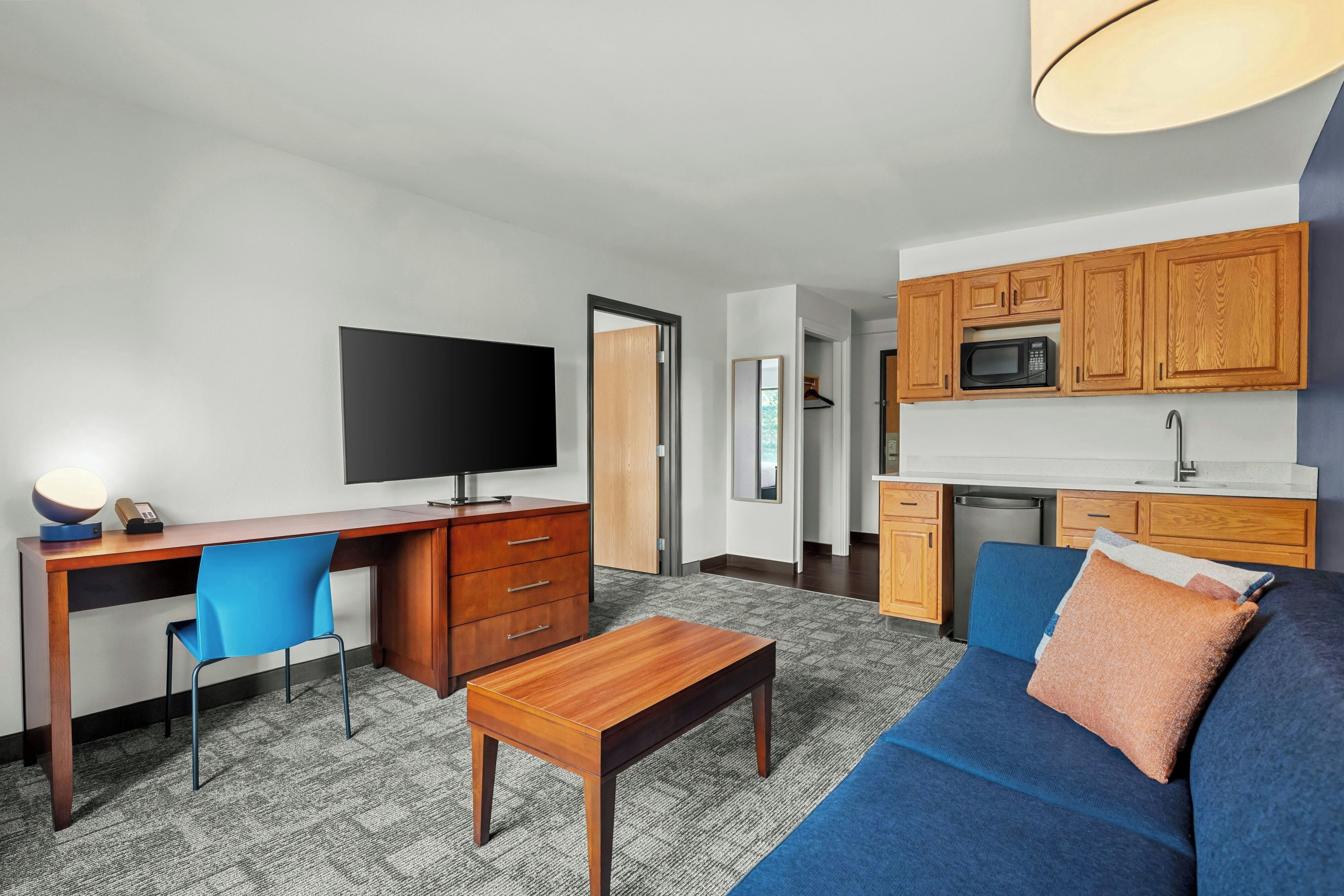 A modern living room with a flat-screen TV, wooden desk, blue sofa, and a kitchen area with wooden cabinets and a microwave.