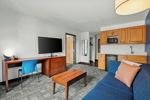 Een moderne woonkamer met een flatscreen televisie, een houten bureau, een blauwe bank en een keuken met houten kastjes en een magnetron.