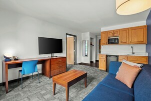 Una sala de estar moderna con televisor de pantalla plana, escritorio de madera, sofá azul y una zona de cocina con armarios de madera y un microondas.