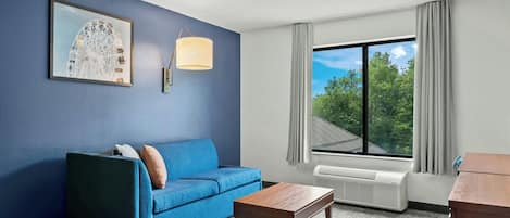 A hotel room with a blue sofa, a small wooden table, a framed picture on the wall, a window with a view of trees, and a ceiling-mounted lamp.