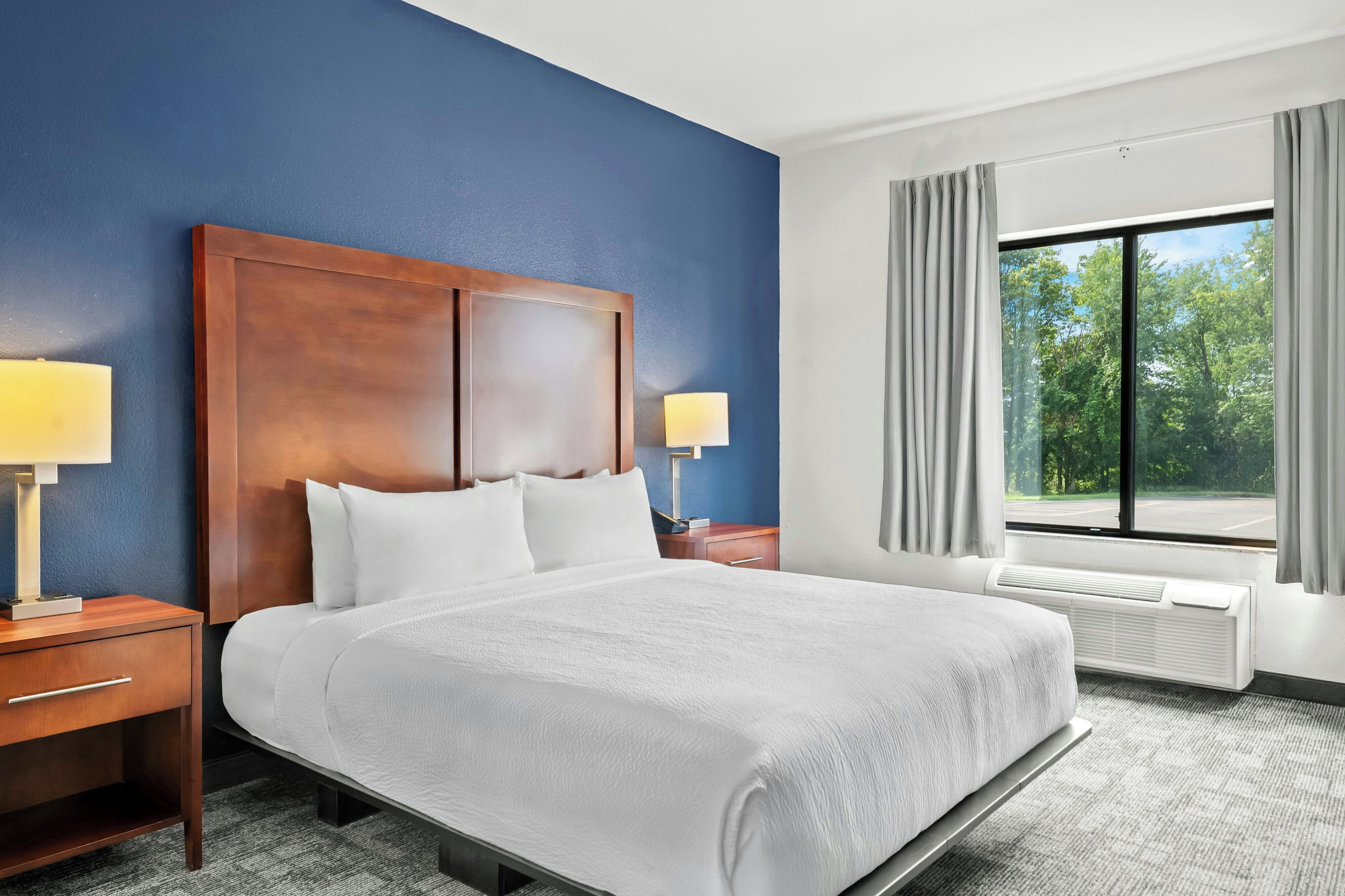 Una camera d'albergo con un letto grande, due lampade ai lati del letto, una testiera in legno, un comodino, una finestra con tende e un condizionatore d'aria.