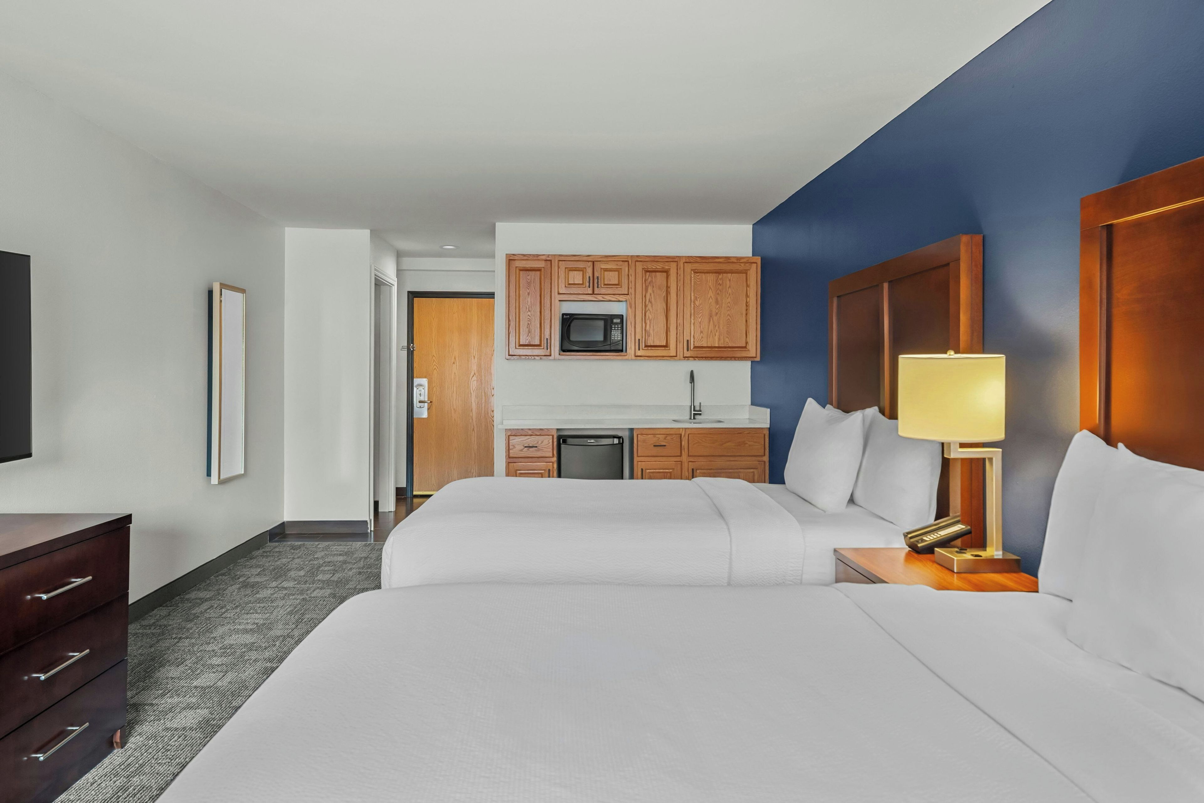 A hotel room with a bed, bedside table, lamp, and a kitchenette with wooden cabinets and a microwave.