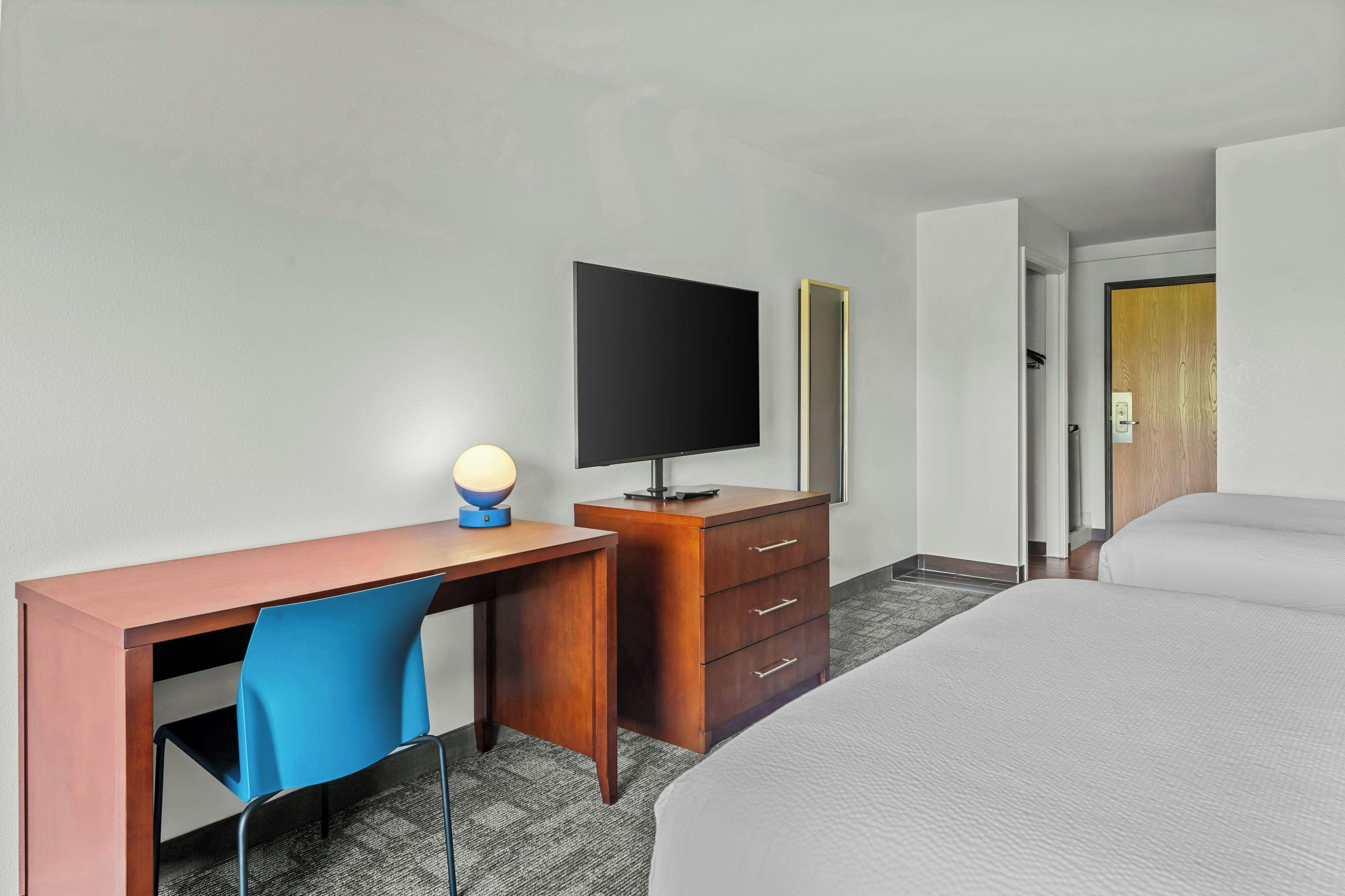 Ett hotellrum med en säng, ett skrivbord med en stol, en tv, en byrå och en spegel.