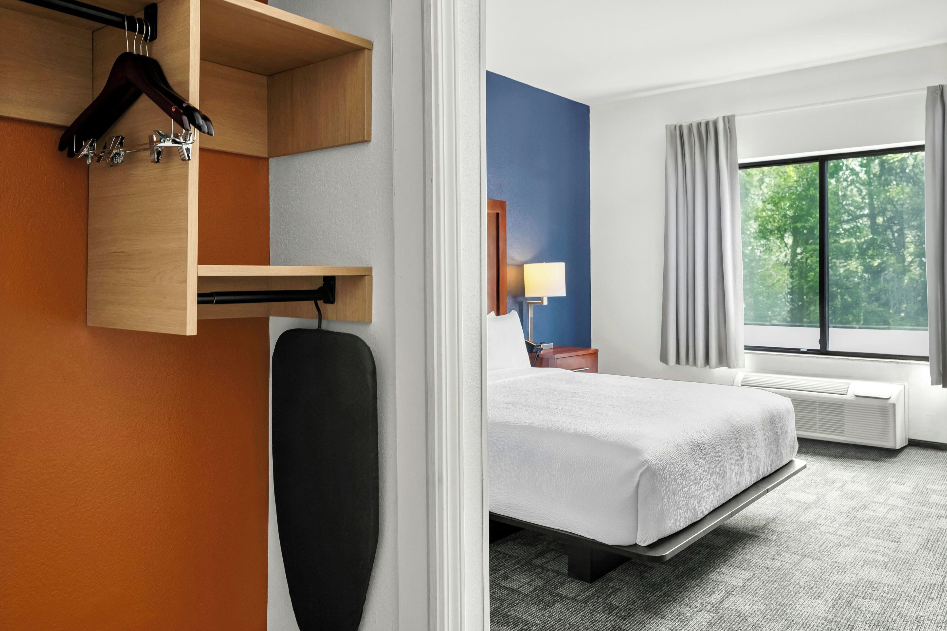 A hotel room with a bed, a bedside table with a lamp, a window with curtains, and a wall-mounted heater.