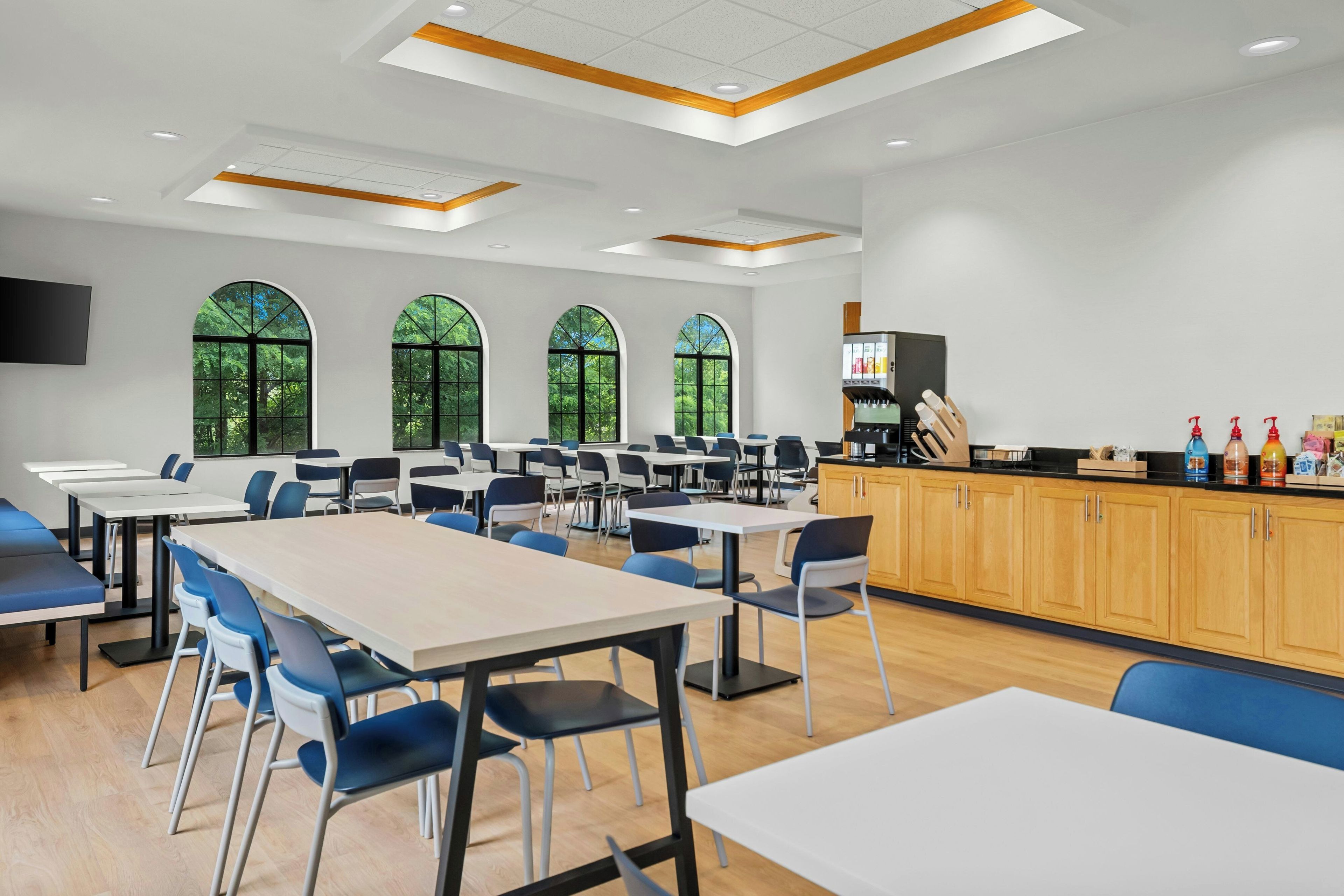 Un vaste espace dédié à la salle à manger, avec de nombreuses tables et chaises, de grandes fenêtres offrant une vue sur la verdure, et un comptoir où sont exposés divers articles.