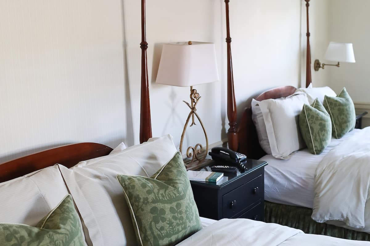 A hotel room with two beds, each with white bedding and green patterned pillows. There is a nightstand with a lamp and a telephone between the beds.