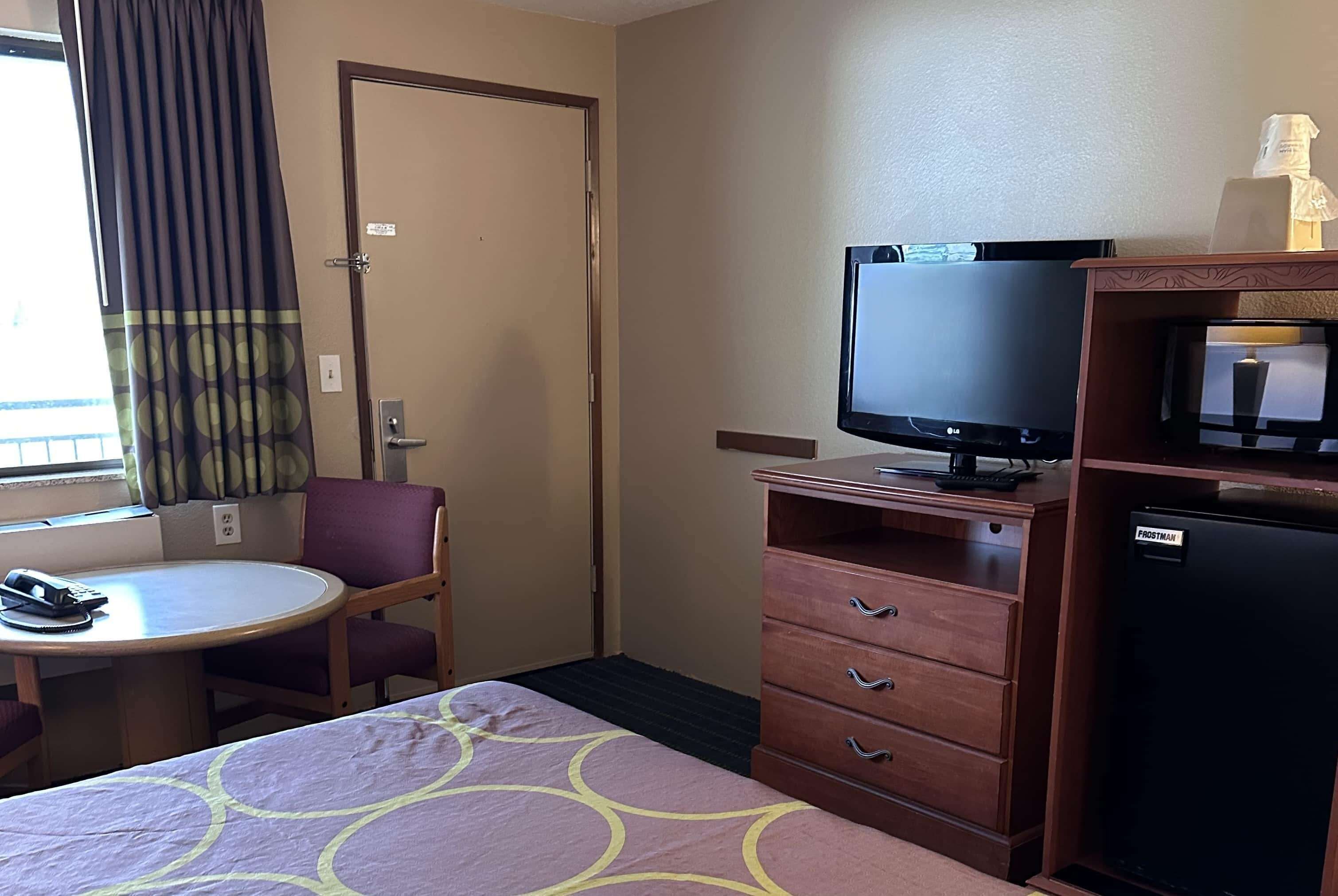 A hotel room with a bed, a desk with a chair, a television, a refrigerator, and a door.