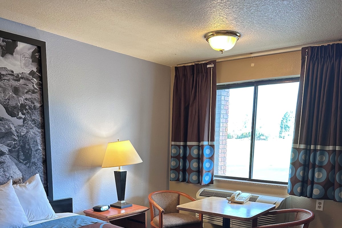 A hotel room with a large bed, a desk with a chair, a lamp, and a window with curtains.