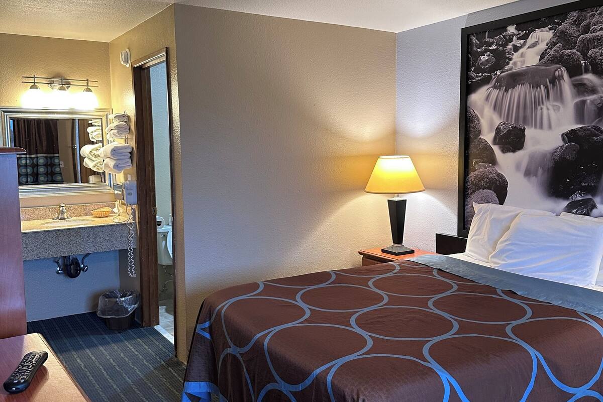 A hotel room with a bed, a desk, a lamp, a bathroom with a sink and mirror, and a large wall painting.