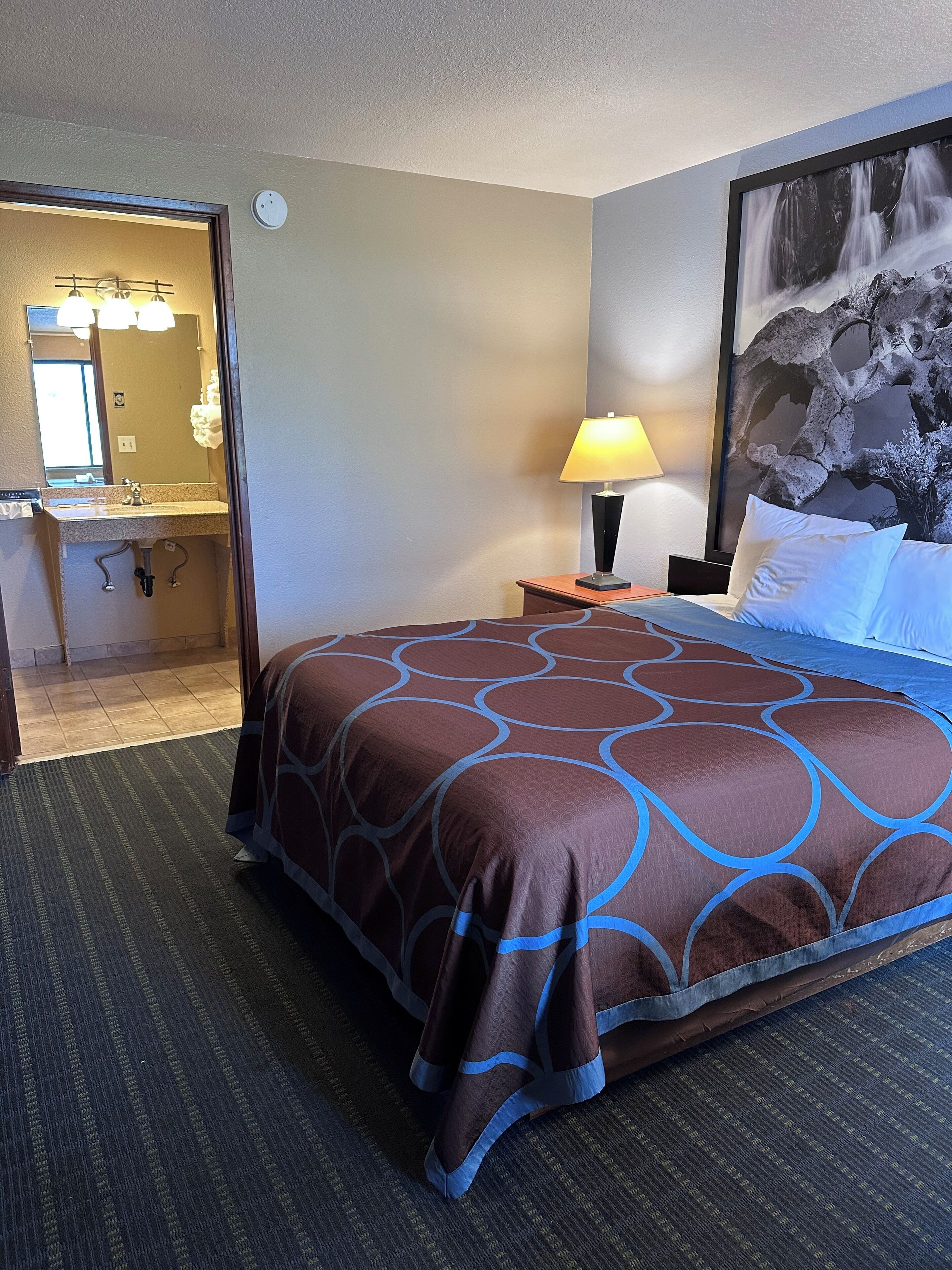 Habitación de hotel con cama, mesita de noche con lámpara, un cuadro grande en la pared y un baño visible a través de una puerta abierta.