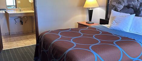 Habitación de hotel con cama, mesita de noche con lámpara, un cuadro grande en la pared y un baño visible a través de una puerta abierta.