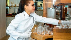 Une personne vêtue d’un uniforme blanc, qui sert des boissons dans un bar doté d’un comptoir en bois et d’un distributeur en métal.