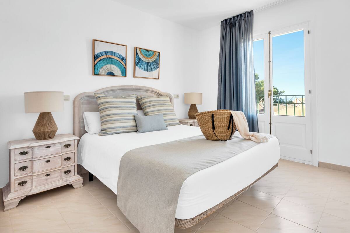 Um quarto com uma cama, duas mesinhas de cabeceira, um criado-mudo, uma janela grande com cortinas e duas obras de arte penduradas na parede.