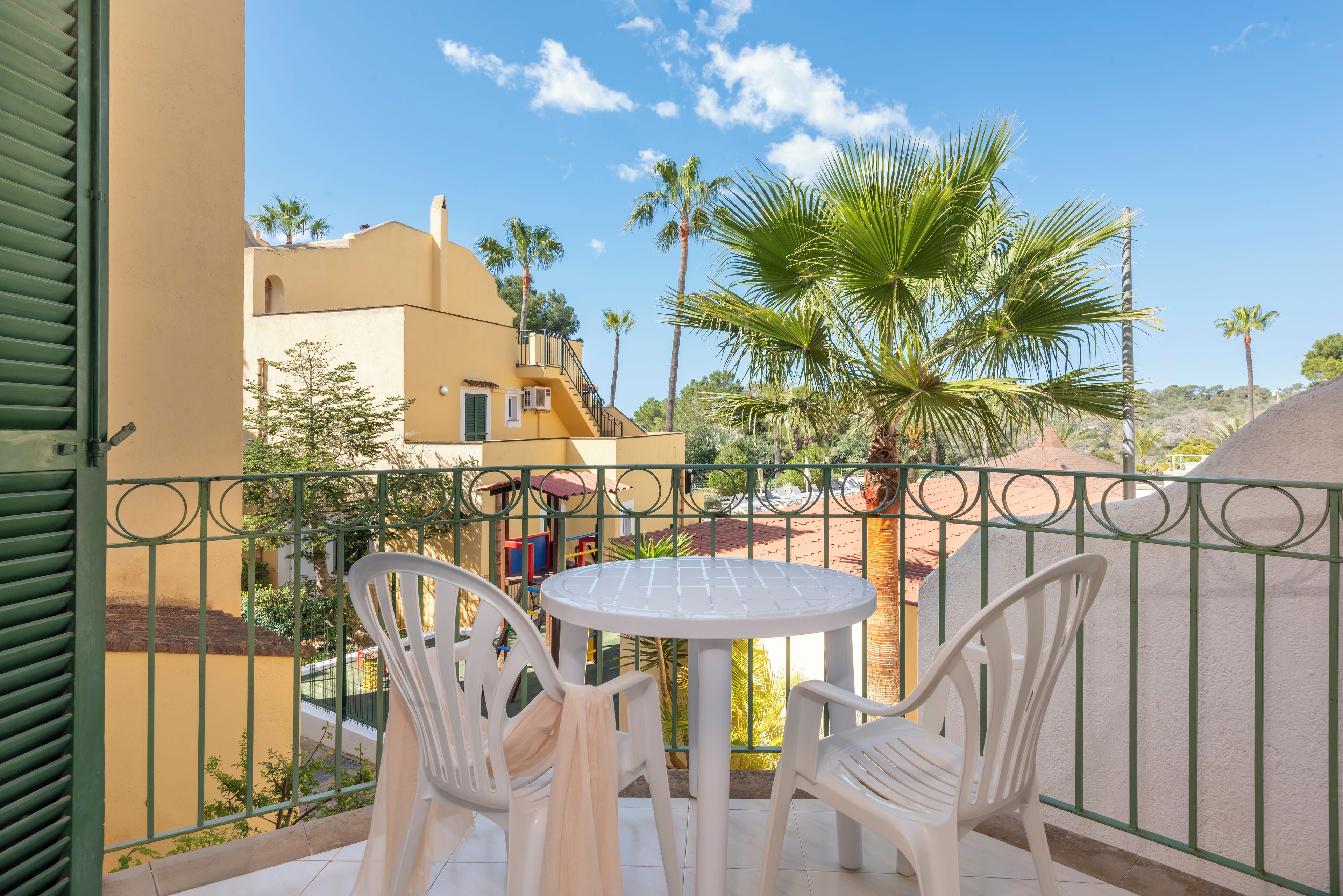 Un balcón con dos sillas de plástico blanco y una mesa redonda pequeña, con vista a un patio con palmeras y un edificio de paredes amarillas.