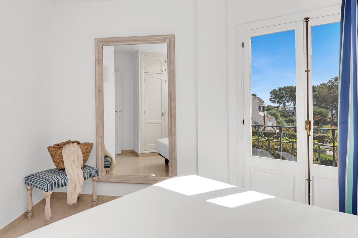 Quarto com um espelho grande, uma cama, um banco com almofada e vista para o exterior através de uma porta.