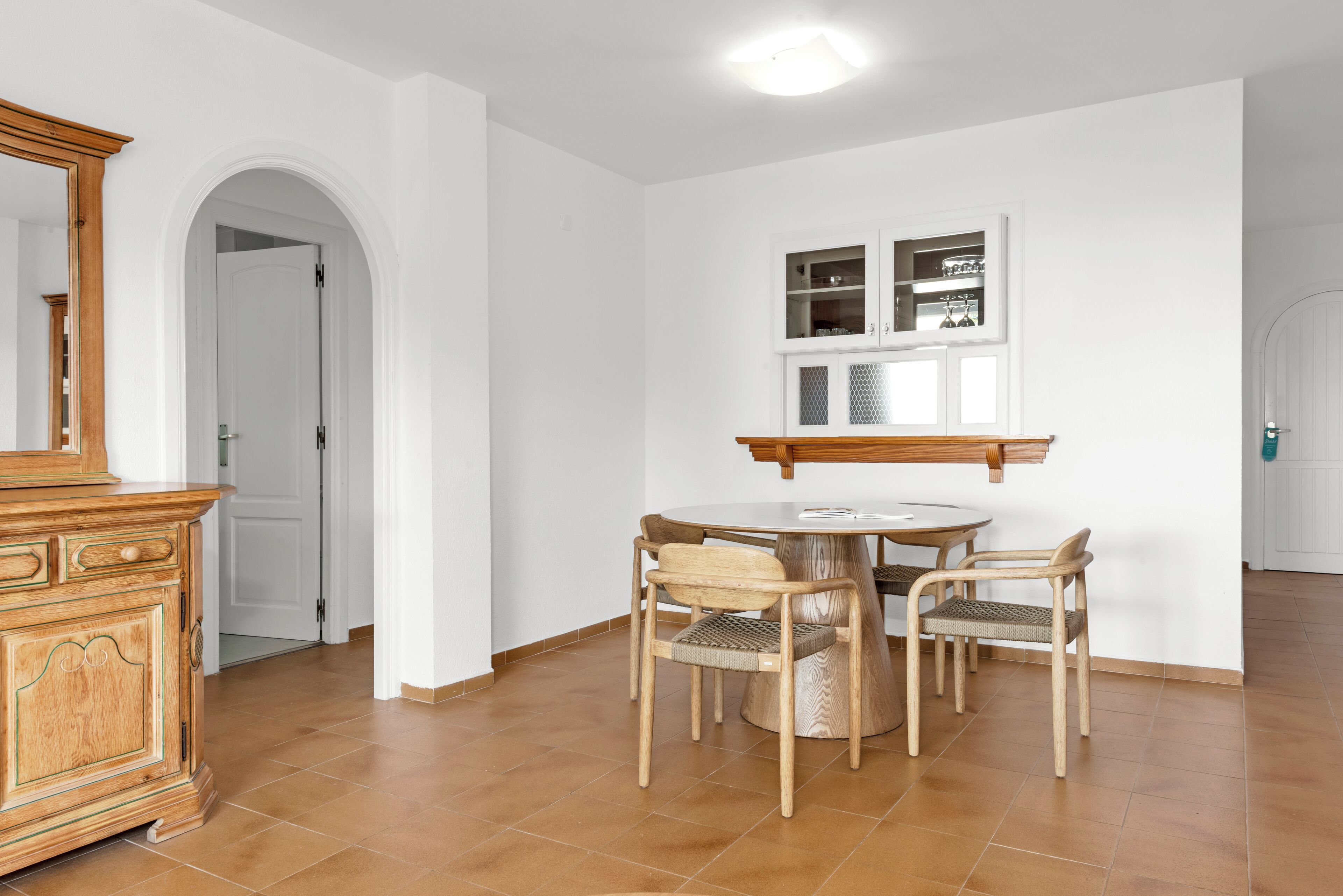 Un coin repas comprenant une table ronde, quatre chaises en bois, un buffet en bois et un mur blanc avec une fenêtre située au-dessus du buffet.