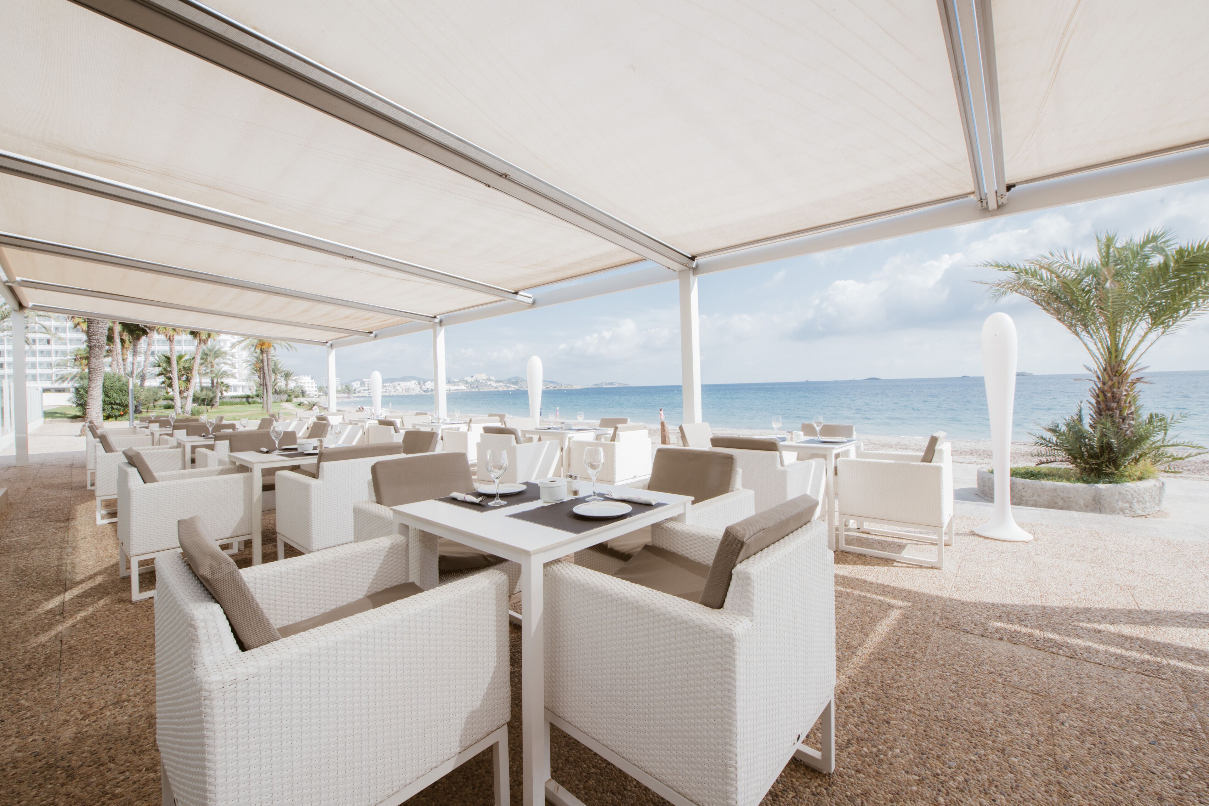An outdoor dining area with white wicker furniture, tables, and chairs, overlooking a body of water.