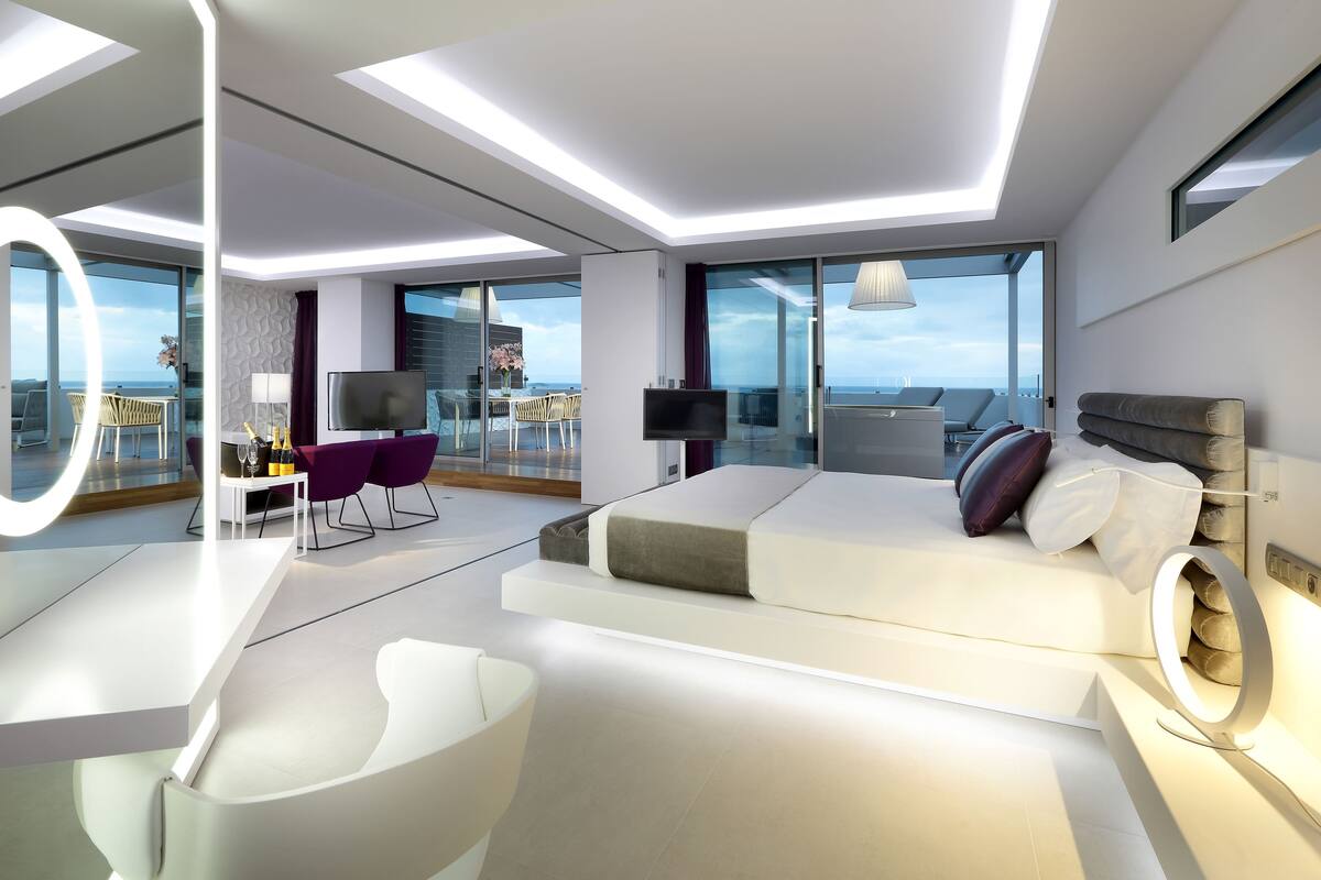 A modern hotel room with a large bed, a desk with a chair, a TV, and a balcony with a view of the sea.