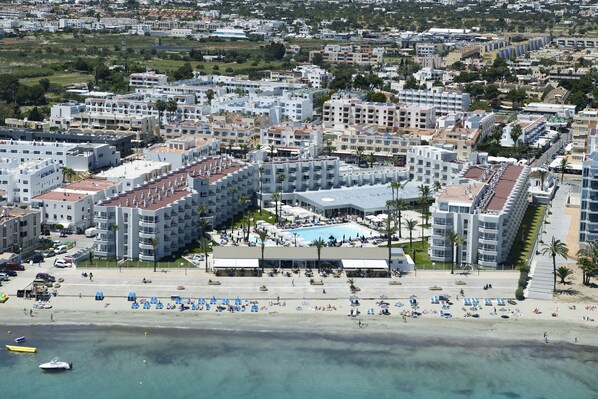 Un hotel frente al mar con varias edificaciones, piscina y zona de playa con tumbonas.