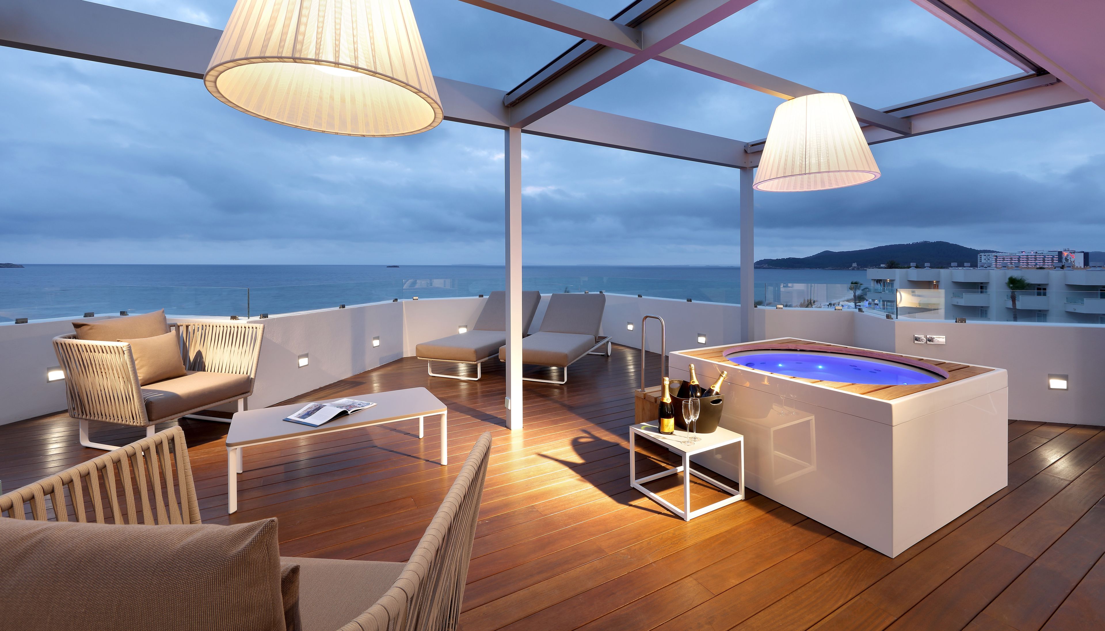 A modern outdoor deck with wooden flooring, lounge chairs, a hot tub, and a view of the sea.