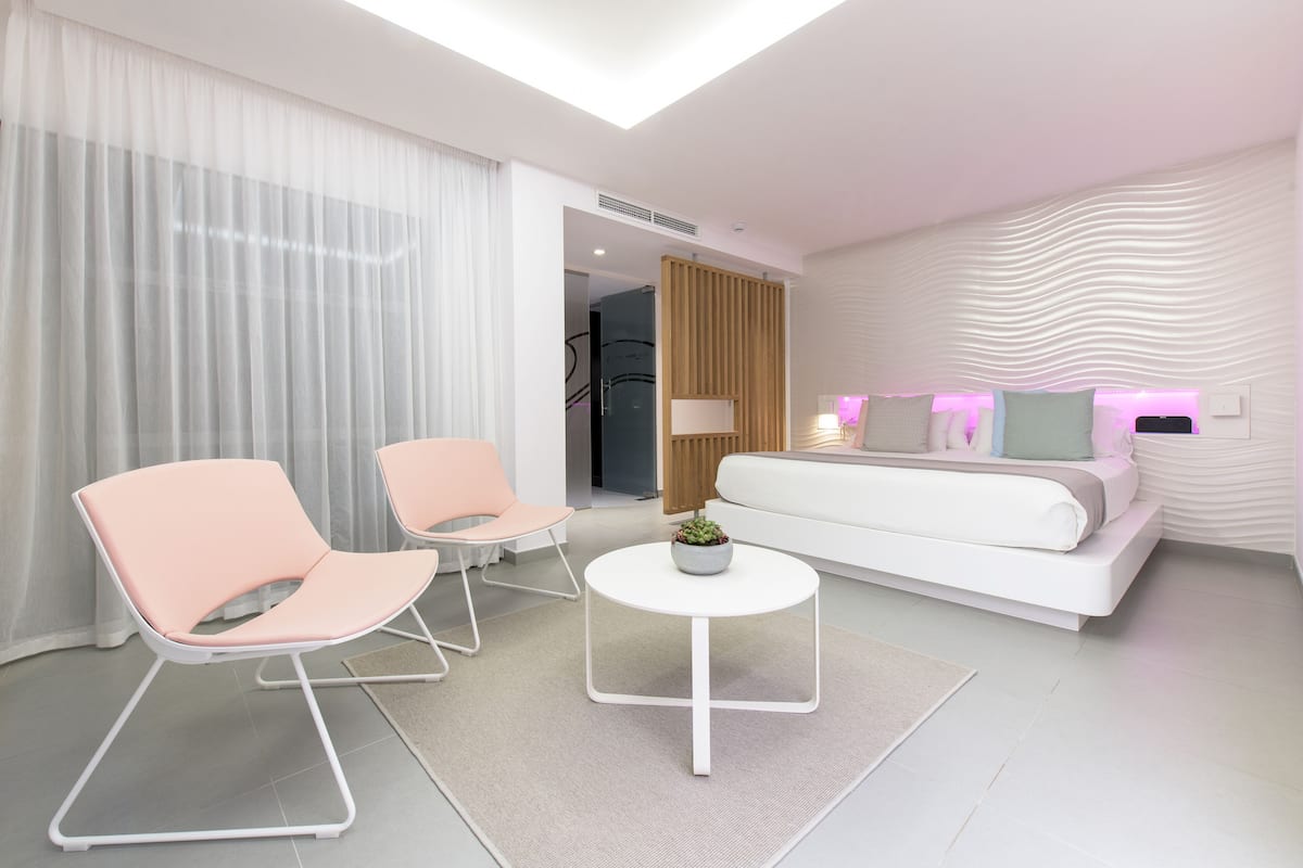 A modern hotel room with a white sofa, two pink chairs, a small round table with a plant, and a large window with sheer curtains.