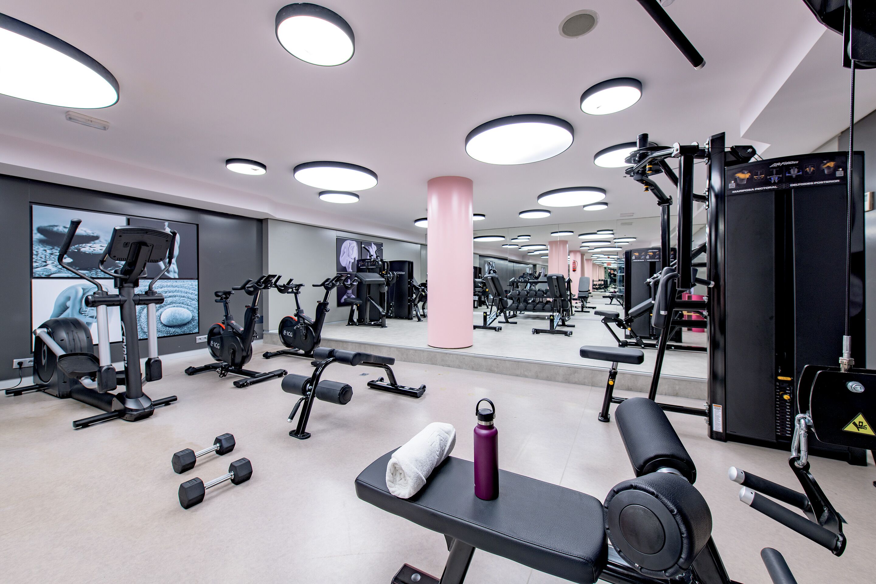 Ein Fitnessraum mit verschiedenen Trainingsgeräten, einer Bank, Kurzhanteln und einer Wasserflasche.