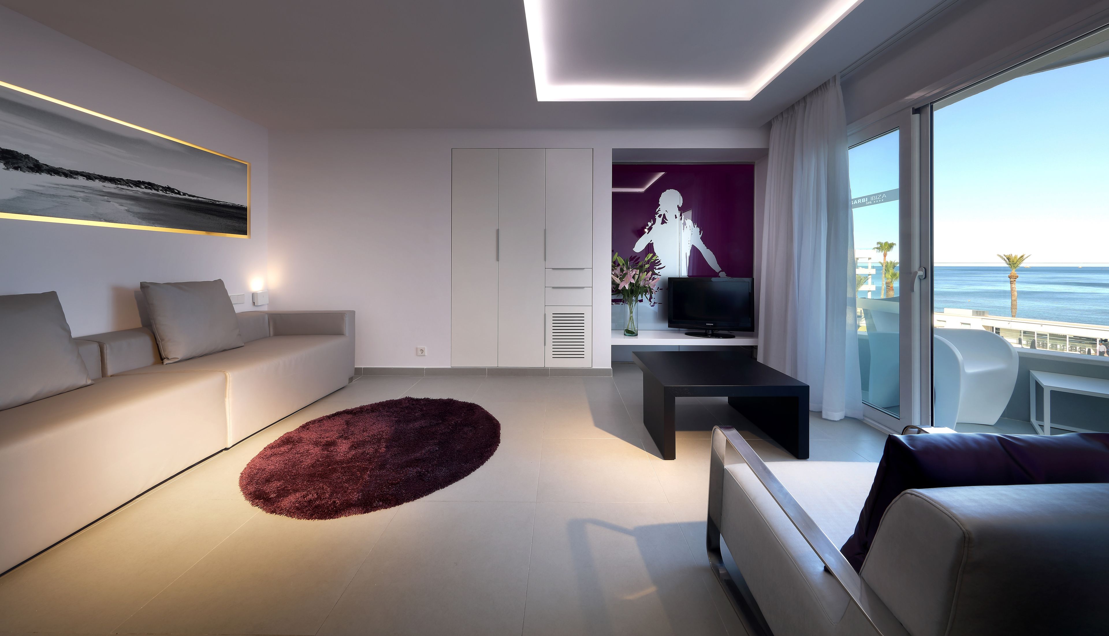 Una sala de estar moderna con un sofá, una mesa de centro y un televisor. Se aprecia una ventana grande con vista al mar.