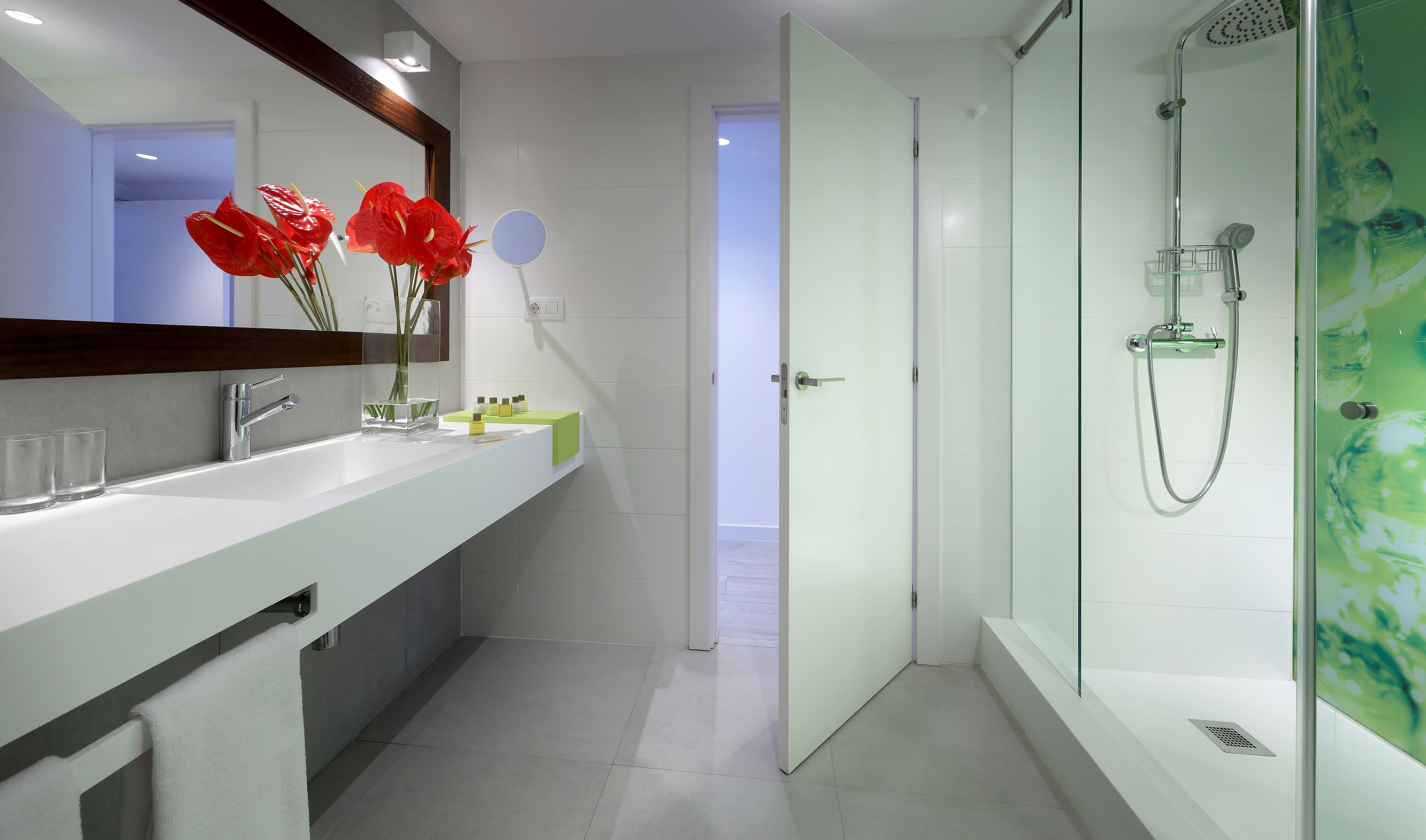 A modern bathroom with a large mirror, a sink with two clear glasses, a vase with red flowers, and a shower area with a glass door.