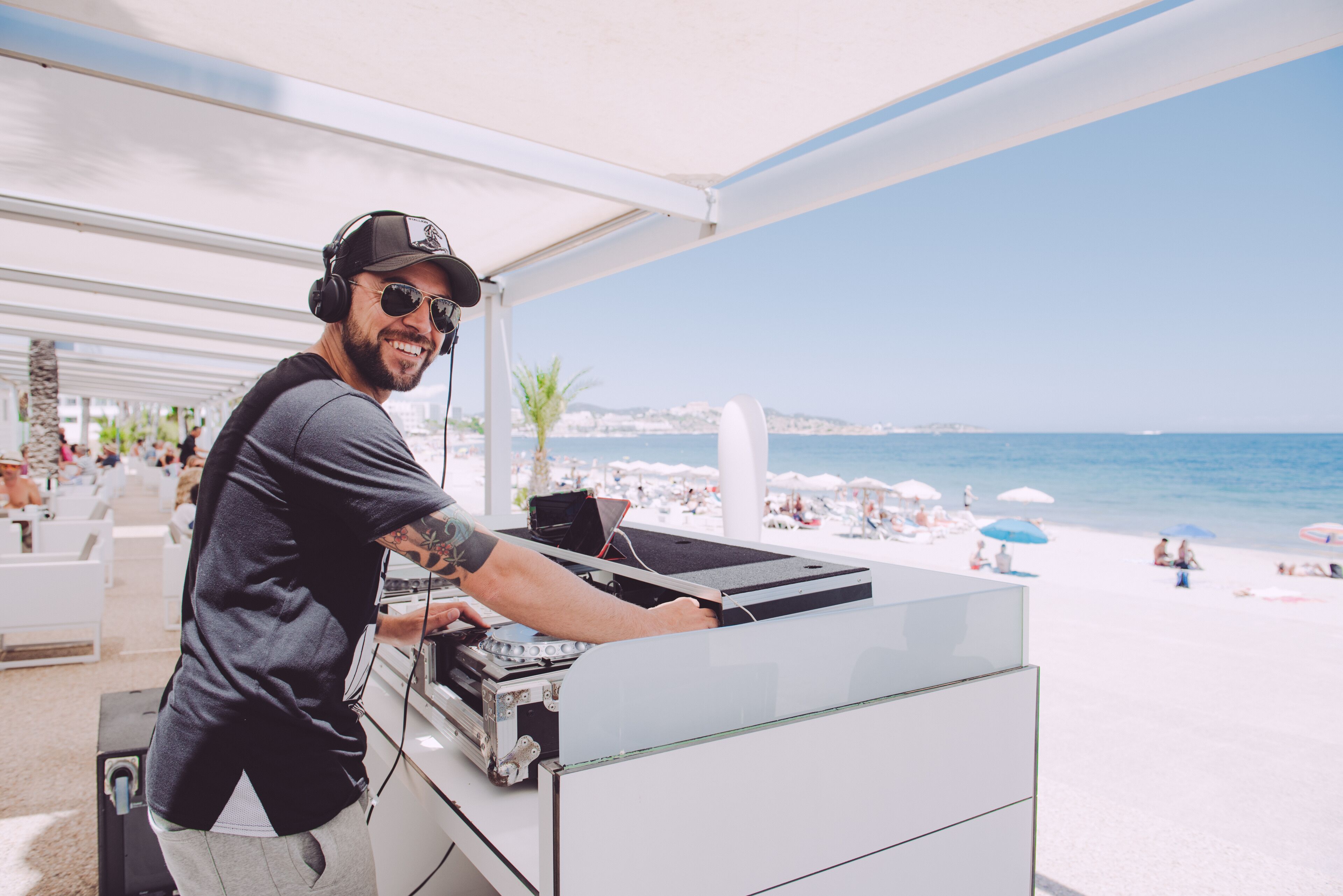 Een DJ-kraam met uitzicht op zee, witte buitenmeubels en parasols.