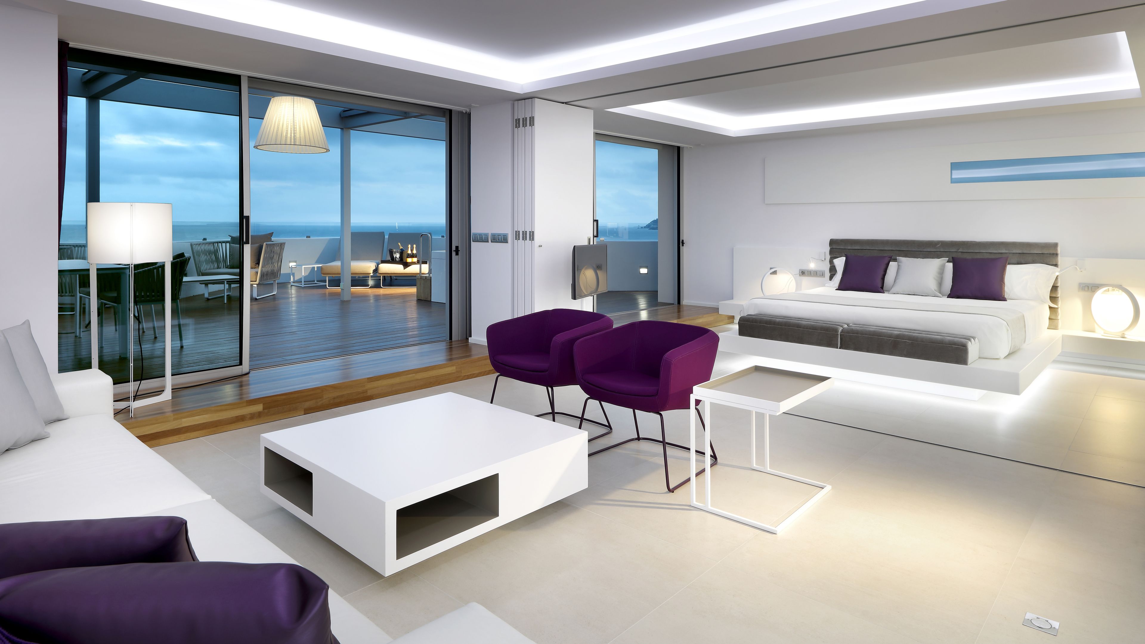 Una habitación de hotel moderna con un amplio ventanal corredero que da a un balcón, una cama con cabecera, una mesa de centro blanca y sillones morados.