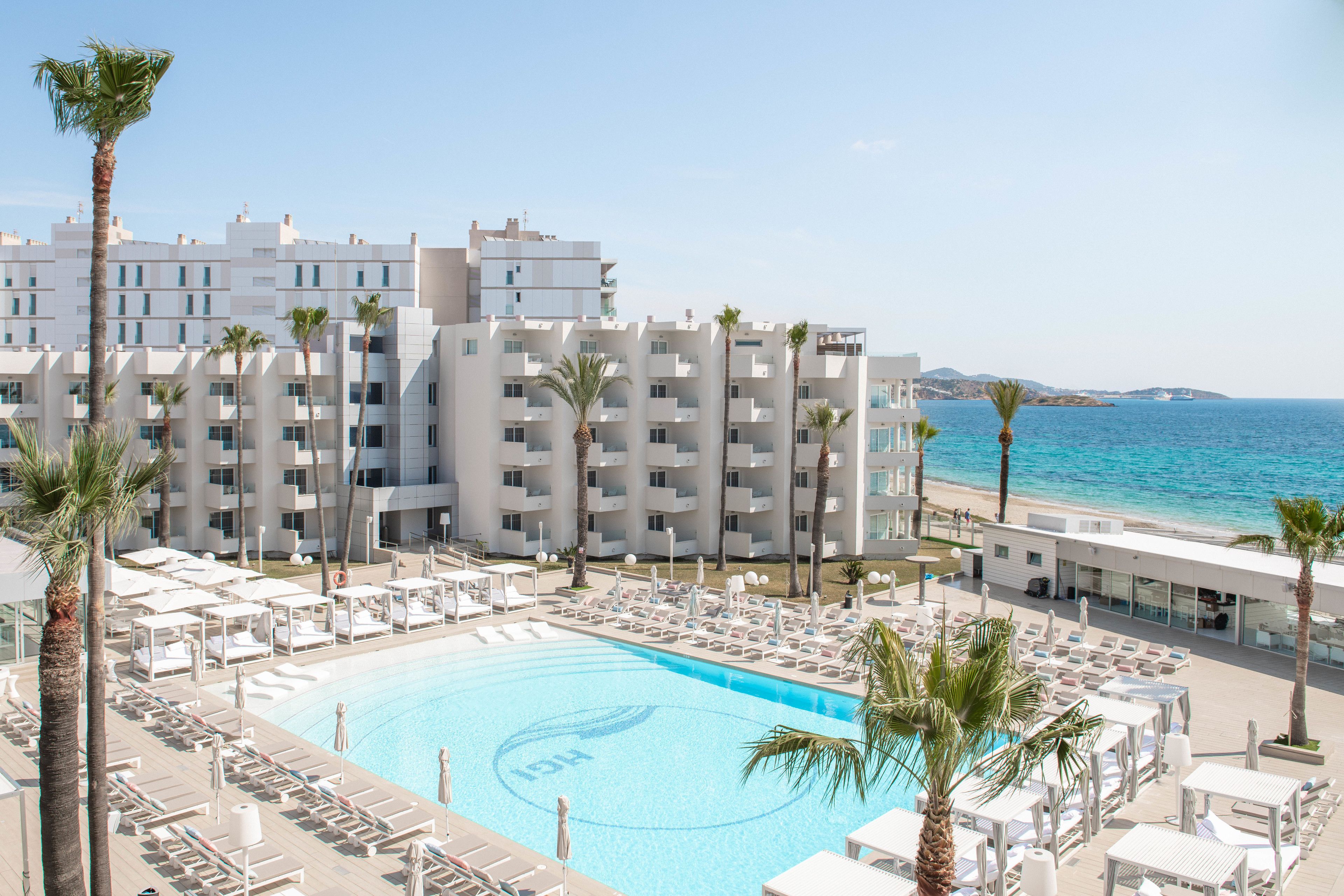 Zona de piscina de hotel con sillones reclinables y sombrillas, una piscina azul cristalina y vistas al mar.