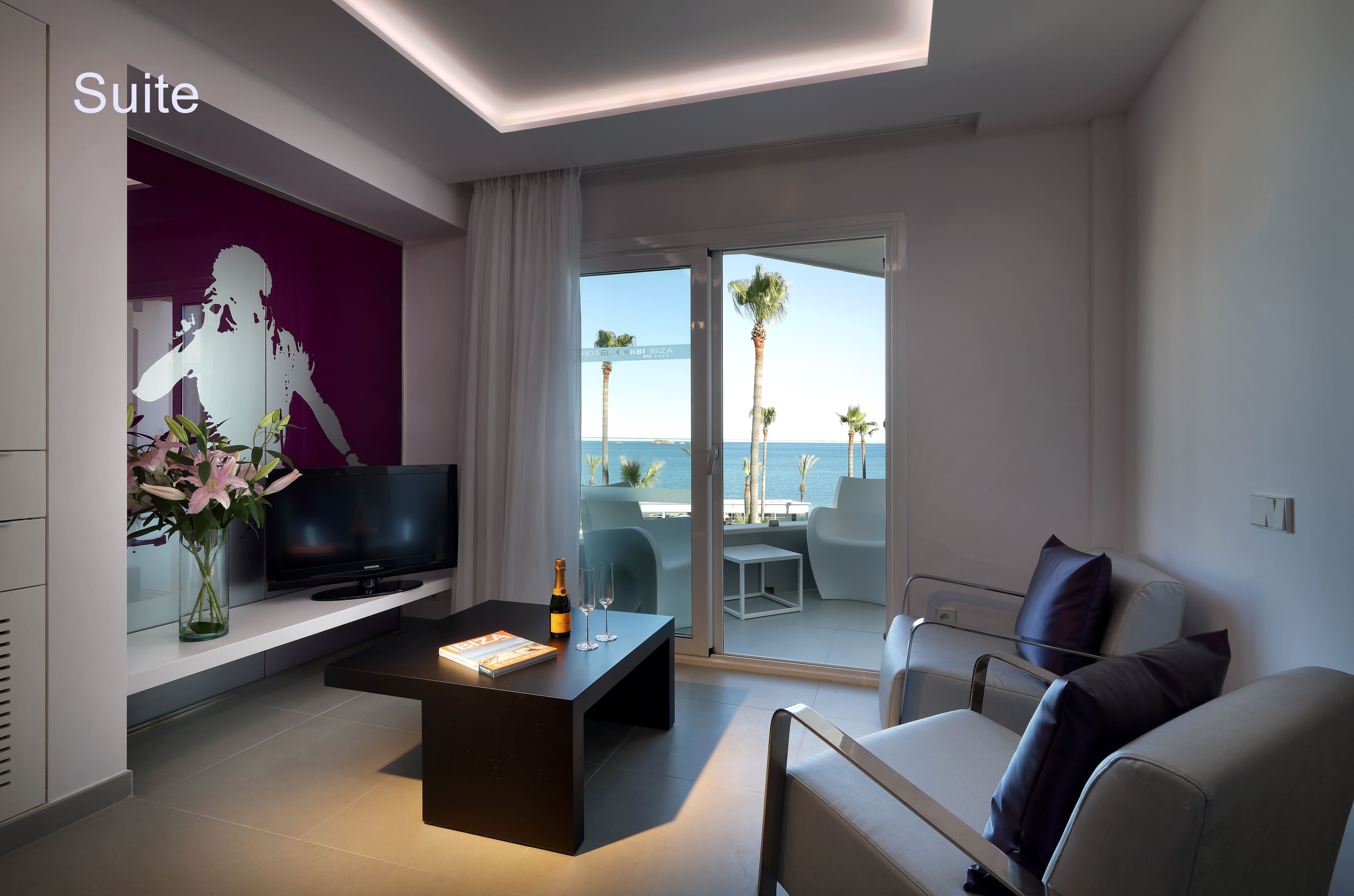 A modern suite with a flat-screen TV, a coffee table, and a sofa. It features a large sliding glass door leading to a balcony with a view of the sea.