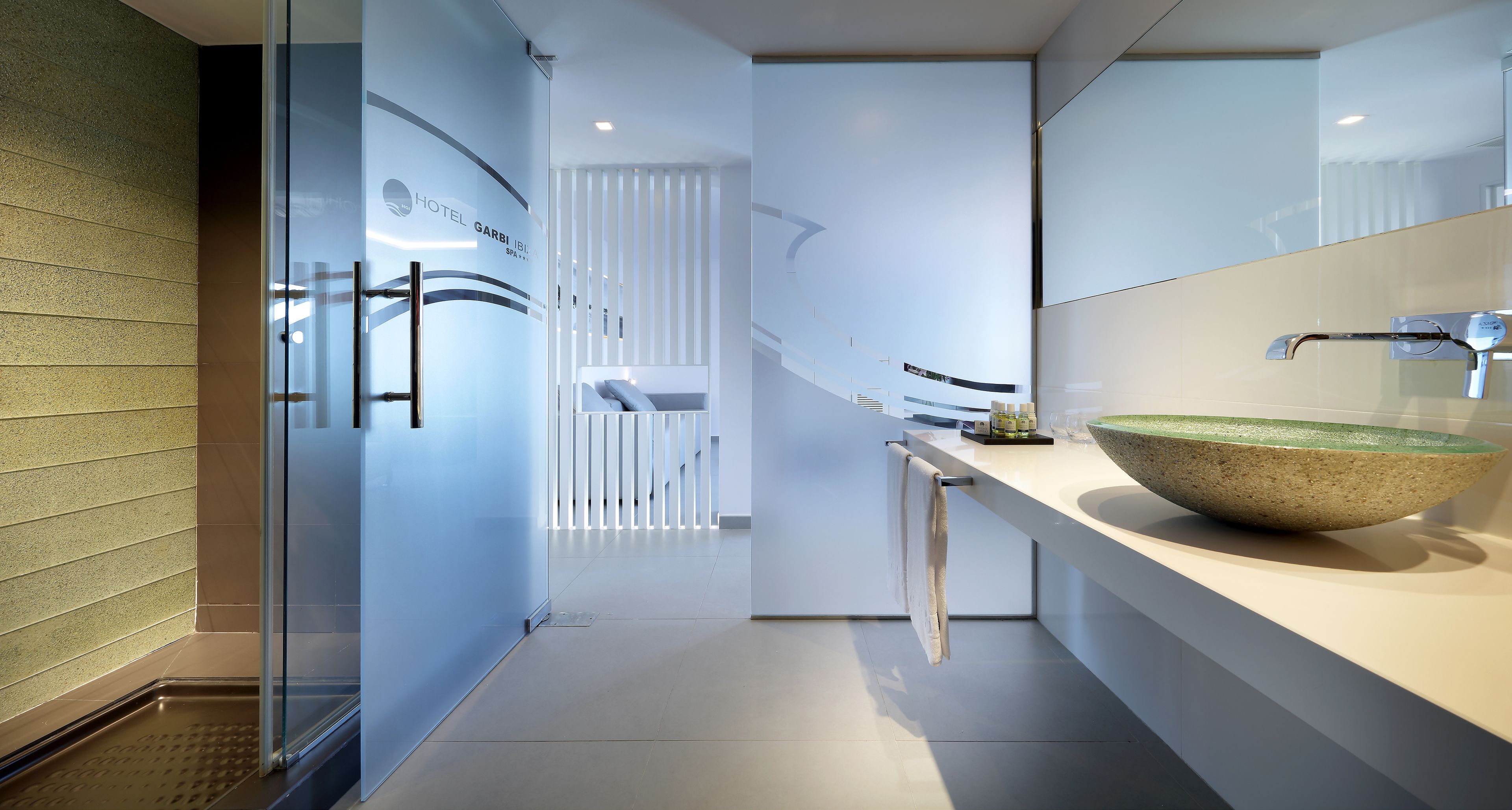 A modern hotel bathroom with a large glass door, a white countertop, and a large bowl sink.
