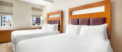 A hotel room with two beds, wooden headboards, white bedding, and large windows with city views.
