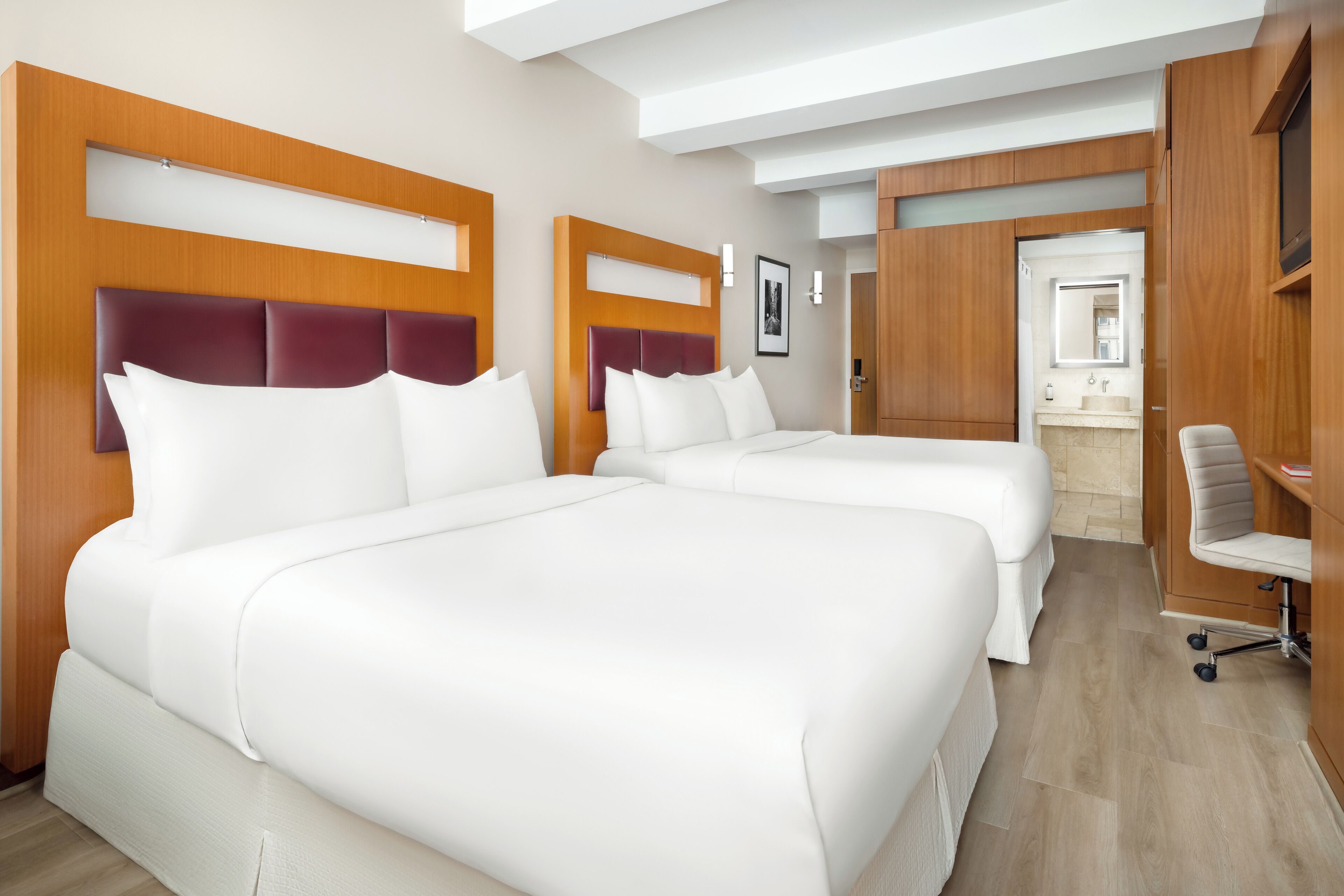 A hotel room with two large beds, a wooden headboard, a desk, and a bathroom with a sink and mirror.